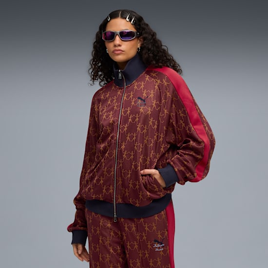 Un modello con occhiali da sole posa in giacca bordeaux con zip e pantaloni abbinati su sfondo grigio.