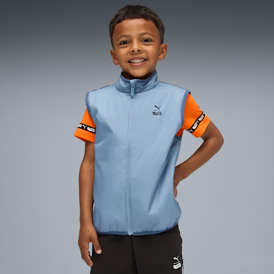 A smiling child poses against grey, wearing a blue sleeveless jacket, orange tee, and dark bottoms.