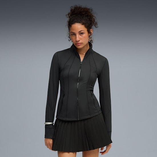 A model with a grey backdrop poses in a black zip jacket and pleated skirt.