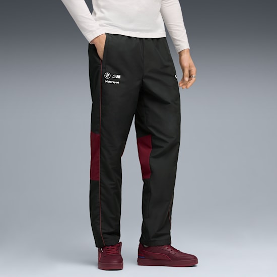 Against grey, a model wears BMW branded black tracksuit bottoms and maroon trainers, hand in pocket.