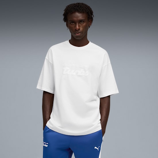 Against grey, a model poses in a white tee and blue track pants, hands in pockets.