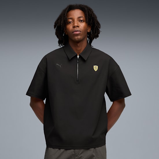 Against grey, a model poses wearing a black, short-sleeved, zipped polo shirt.