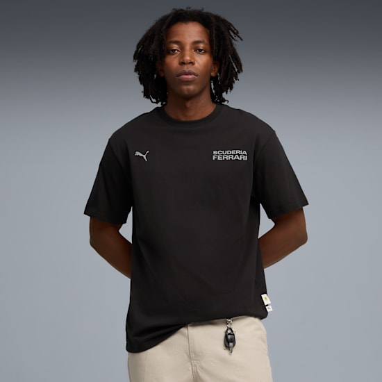 Su uno sfondo grigio, un modello posa indossando una t-shirt nera e pantaloni beige.