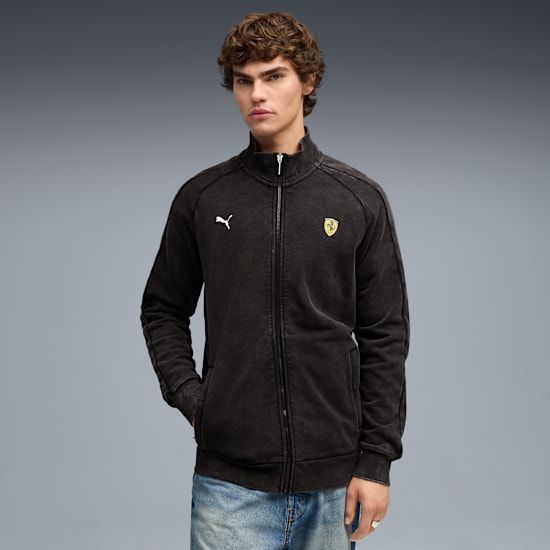 Tegen grijs draagt een model een donker jack met rits, logo's en blauwe jeans.