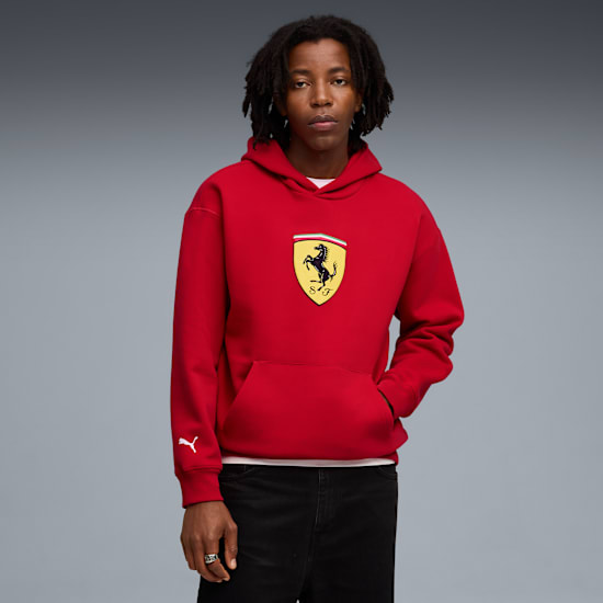 Vor grauem Hintergrund trägt ein Model einen roten Hoodie mit Ferrari-Logo und dunkle Hosen.