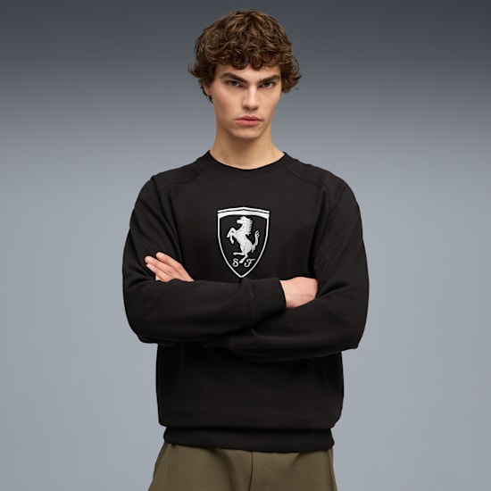 A model with crossed arms wears a black top with a white emblem against a grey backdrop.