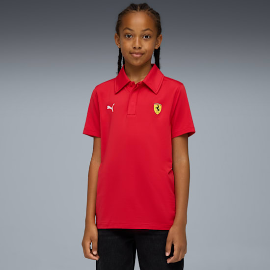 A model with braids poses on grey wearing a red polo shirt with logos and black trousers.