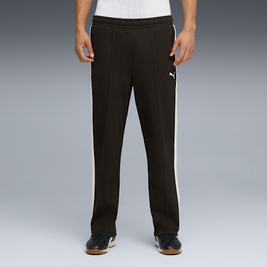 Against grey, a model displays black track pants with a white stripe and dark trainers.