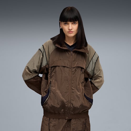 Against grey, a model poses in a brown jacket and trousers with hands in pockets.