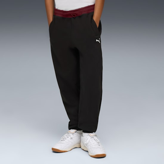 A model stands against grey wearing black joggers and white trainers.