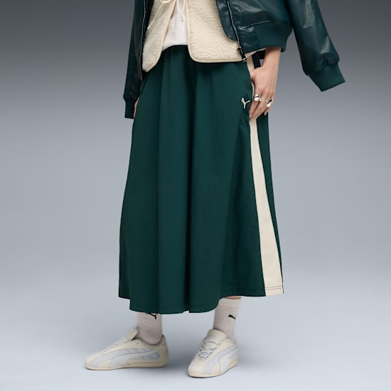 On grey, a model wears a green skirt with stripe, jacket and trainers.