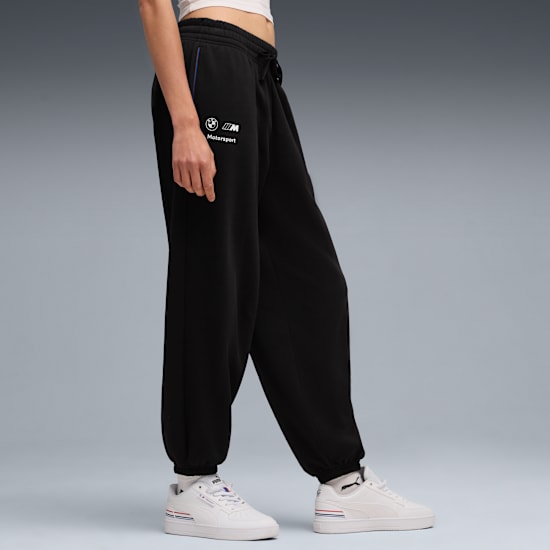A model poses on a grey background wearing black branded trousers and white trainers.