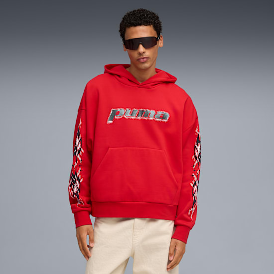 Un modello posa su sfondo grigio, con occhiali da sole, felpa rossa e pantaloni beige.