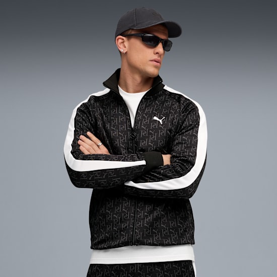 A model with crossed arms wears a patterned jacket, cap and sunglasses against a grey backdrop.