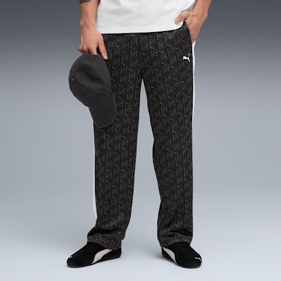 A model poses against grey, wearing patterned black trousers, trainers, and holding a cap.