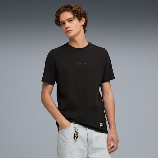 Na szarym tle model pozuje w czarnym t-shircie i jasnoniebieskich dżinsach, z ręką w kieszeni.