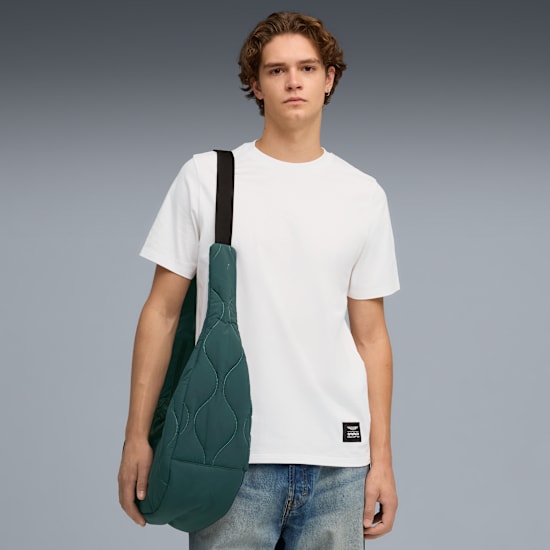 Na szarym tle model ma na sobie białą koszulkę, dżinsy i pikowaną zieloną torbę z czarnym paskiem.