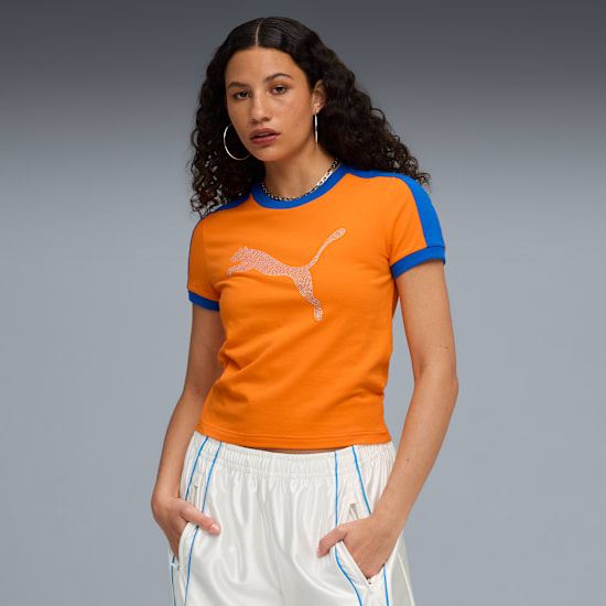 Against grey, a model poses with hands in pockets, wearing an orange and blue t-shirt and white trousers.