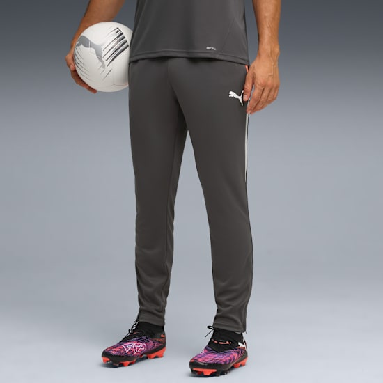 A model holds a football against a grey backdrop, wearing grey Puma sportswear and football boots.