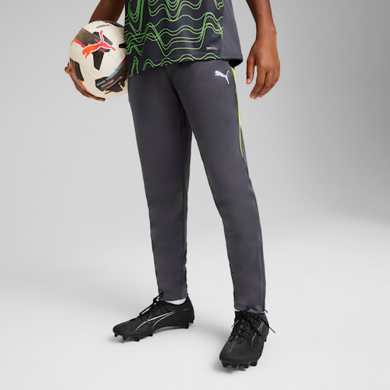 Sur fond gris, un modèle tient un ballon, vêtu d'un haut à motifs, d'un pantalon gris et de chaussures de foot noires.