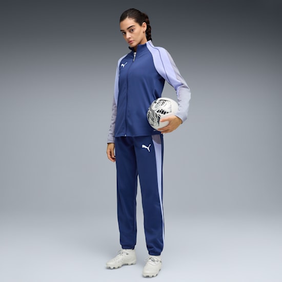On grey, a model holds a football, wearing a blue tracksuit and trainers.