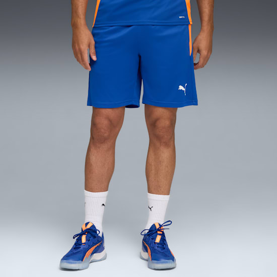 Tegen een grijze achtergrond draagt een model een blauwe short, bijpassende schoenen en witte sokken.