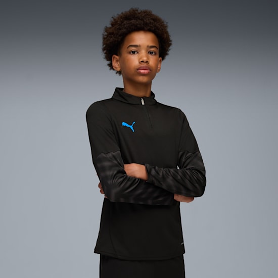 Vor grauem Hintergrund posiert ein Model mit verschränkten Armen in einem schwarzen Oberteil mit blauem Logo.