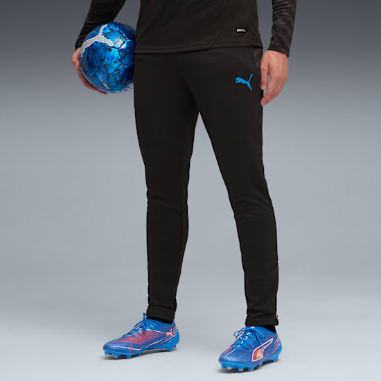 A model against grey holds a football, wearing black tracksuit bottoms and blue football boots.