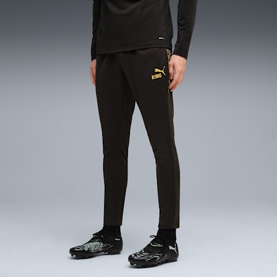 Against grey, a model wears black Puma tracksuit bottoms and football boots.