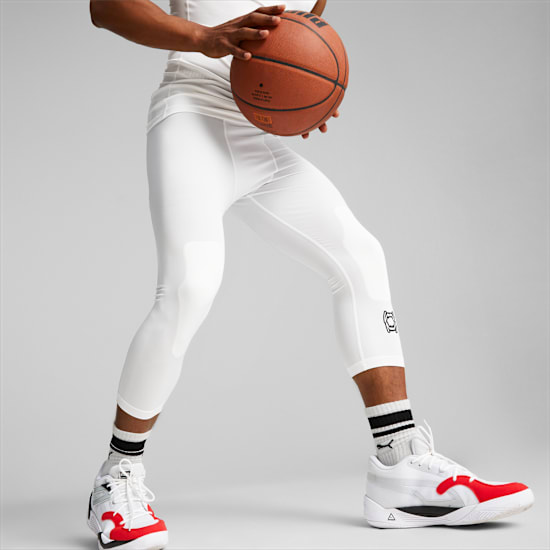 Vor grauem Hintergrund hält ein Model einen Basketball, trägt weiße Leggings und Schuhe.