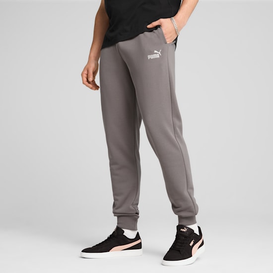 Vor grauem Hintergrund trägt ein Model graue Jogginghosen, ein schwarzes Oberteil und schwarze Turnschuhe.