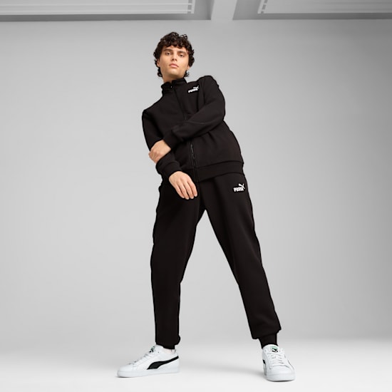 A model poses against a grey backdrop wearing a black tracksuit and white trainers.