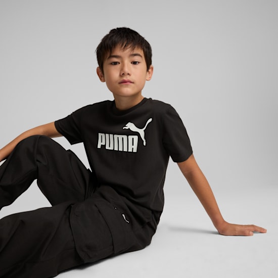 A model in front of a grey background sitting wearing a black Puma t-shirt and black trousers