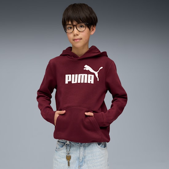 A child with glasses poses in front of a grey background wearing a burgundy Puma hoodie.