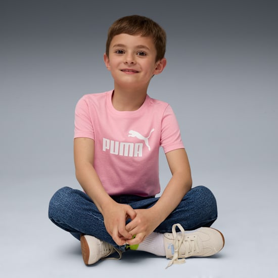 A smiling child sits cross-legged against a grey backdrop, wearing a pink Puma t-shirt and jeans.