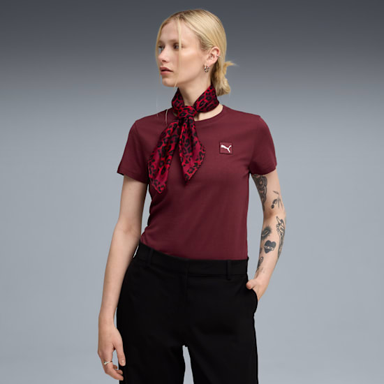Against grey, a model wears a maroon tee, black trousers, and a patterned red scarf.
