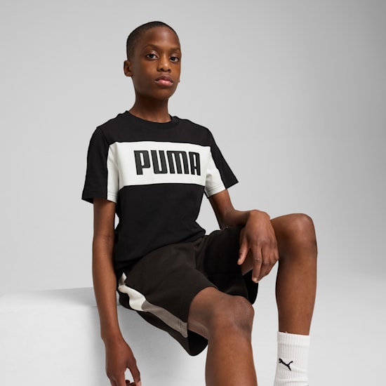 On a grey backdrop, a model poses while wearing a black and white PUMA t-shirt and black shorts.