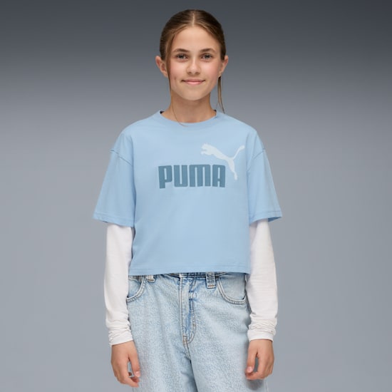 Una modella posa su sfondo grigio, indossando una T-shirt PUMA blu, jeans e una maglia bianca a maniche lunghe.