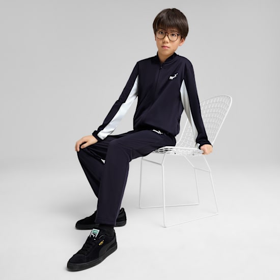Against a grey backdrop, a model sits in a navy tracksuit and black trainers.