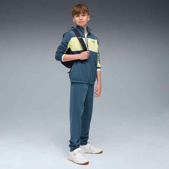 A boy stands against grey, wearing a tracksuit, trainers, and carrying a rucksack.