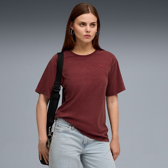 Against grey, a model poses wearing a maroon tee, jeans, and a black shoulder bag.