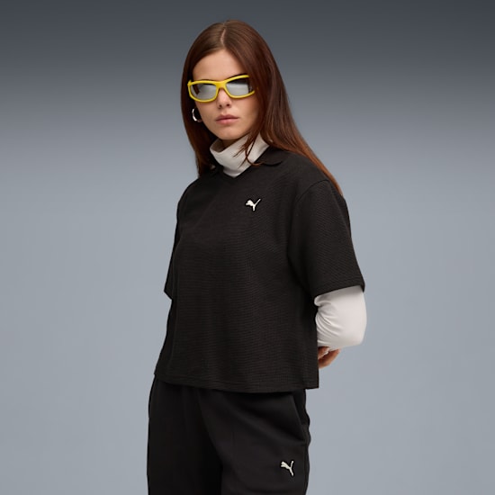 A model with yellow sunglasses poses against a grey backdrop wearing a black top and trousers.