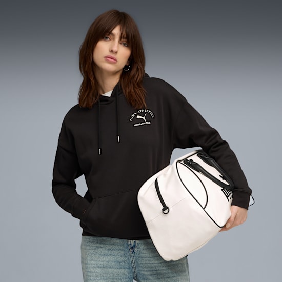 A model holds a white bag, wearing a black hoodie and jeans against a grey backdrop.