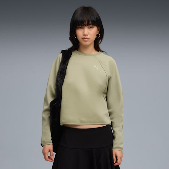 A model with a black bag poses against grey, wearing a green top and black skirt.