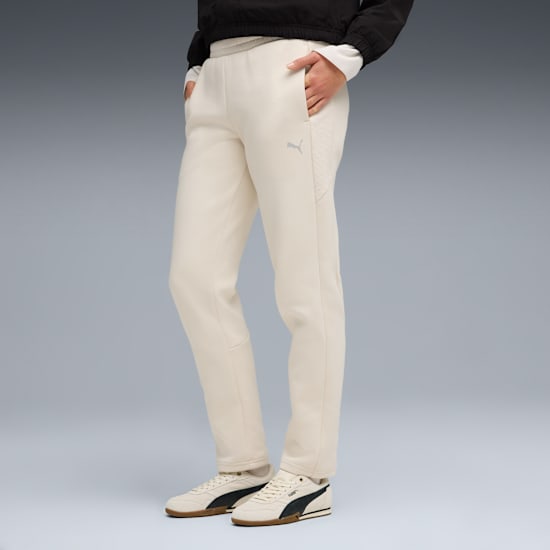 Vor grauem Hintergrund posiert ein Model in cremefarbener Hose und Turnschuhen, die Hand in der Tasche.