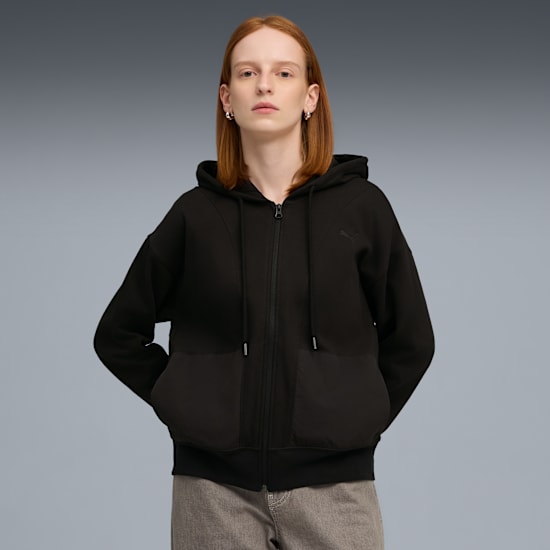 A model with auburn hair poses in a black zip-up hoodie and grey trousers against a grey backdrop.