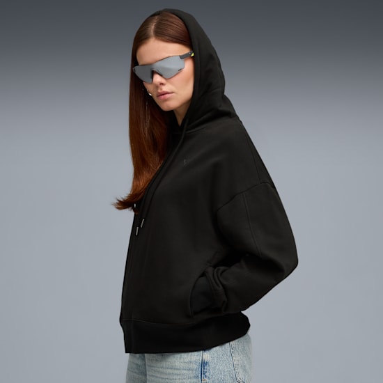 A model with sunglasses poses in front of a grey background wearing a black hoodie and jeans.