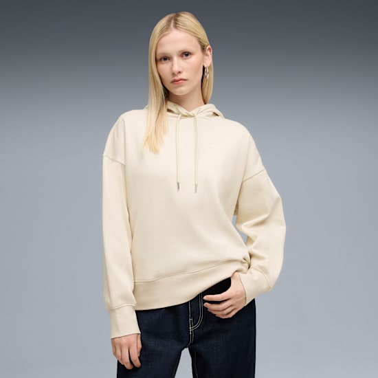 A model against grey wears a cream hoodie and dark jeans, looking at the camera.