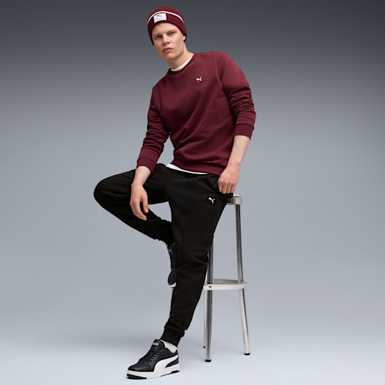 A model on a grey background sits on a stool wearing a burgundy sweatshirt, beanie and black pants.