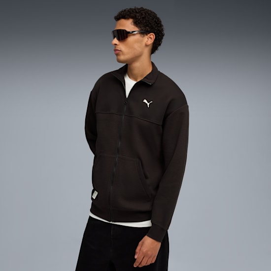 A model with sunglasses poses against a grey backdrop wearing a black jacket and dark trousers.